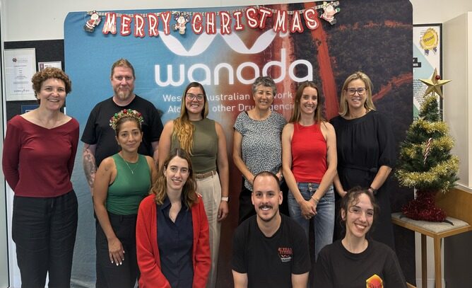 Ten people smiling in front of a media wall with a Merry Christmas banner above and a small Christmas tree to the right.