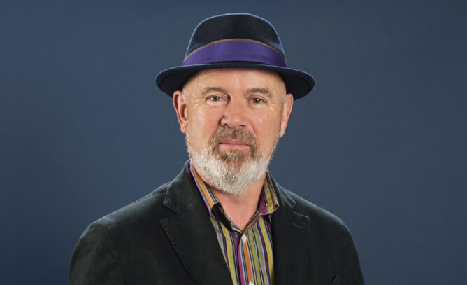 A man with a beard wearing a purple-trimmed fedora hat and black jacket