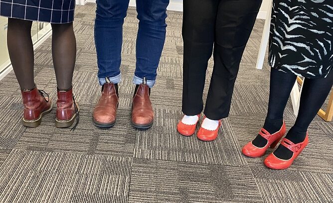 Four pairs of red shoes or boots attached to four pairs of legs. Some footwear has the colour worn off in parts, others are bright red.