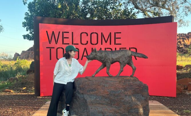 A woman (Christel Barthelemy) looking at a statue of "Red Dog" with a big red sign behind her that reads "Welcome to Dampier"