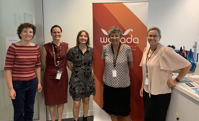 Five women smiling and standing in an office with an orange WANADA banner in the background.