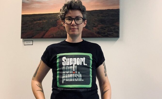A woman with short hair smiling and wearing a black t-shirt with the words 'Support Don't Punish' on it.