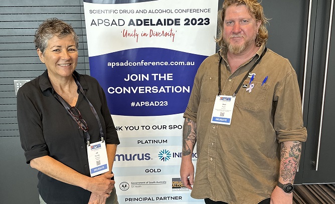 A woman with grey curly hair smiling with a man in a brown shirt with tattoos on his arm standing in front of an APSAD banner.
