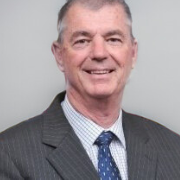 A happy-looking man with short grey hair smiling and wearing a grey jacket, thin-striped white shirt and a patterned blue tie.