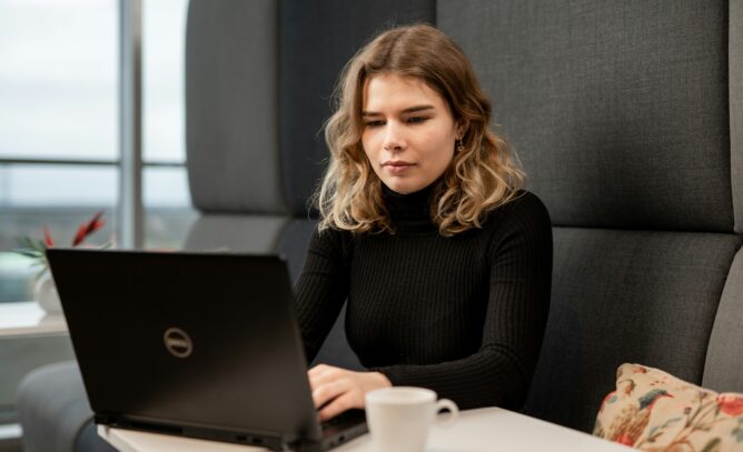A woman with shoulder length hair in a black turtleneck uses a laptop by a window coffee cup at her side