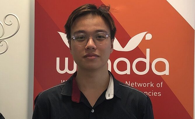 Young Asian man in a grey button-up shirt in front of an orange WANADA banner