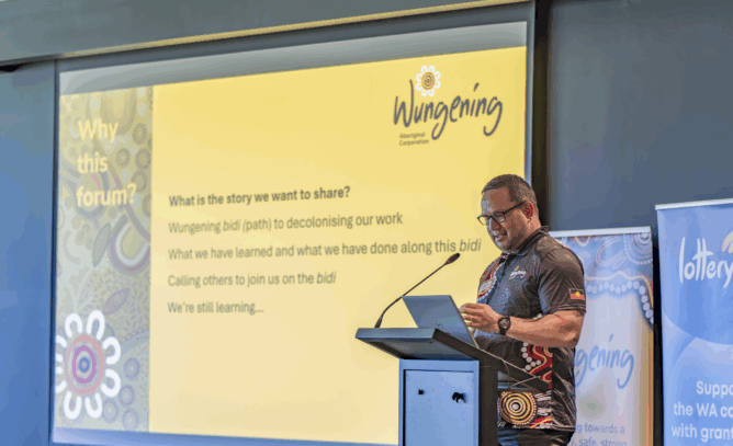 An Aboriginal man stands at a lectern presenting at a forum, reading from notes while speaking into a microphone, with a large yellow presentation slide behind him displaying the Wungening Aboriginal Corporation logo and dot points