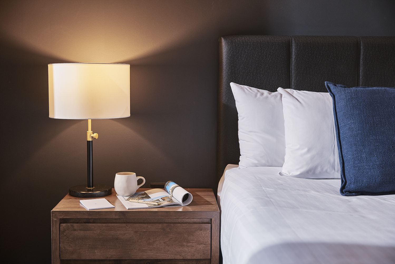 Hotel room with a crisply-made bed with large pillows and a bedside table with a shaded lamp and mug.