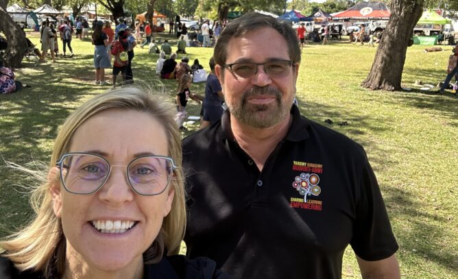 A smiling woman with blonde hair and glasses stands to the left of the frame, in front of a man with brown hair, glasses, and a black polo shirt. They are attending a NAIDOC Week event at a sunny park with clear blue skies in Kununurra.